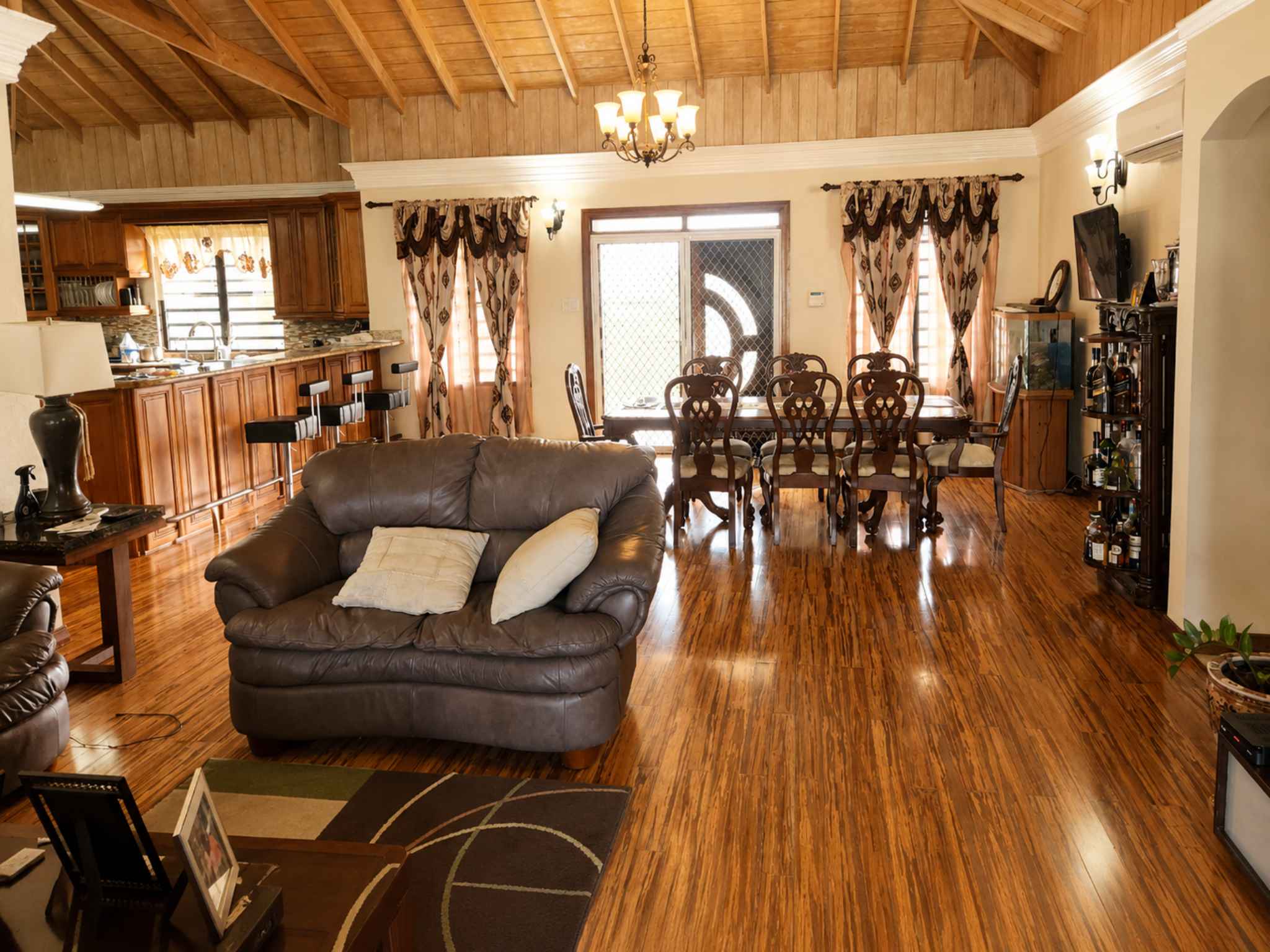 Furnished open-plan living and dining area with timber ceiling, chandelier and kitchen bar beyond
