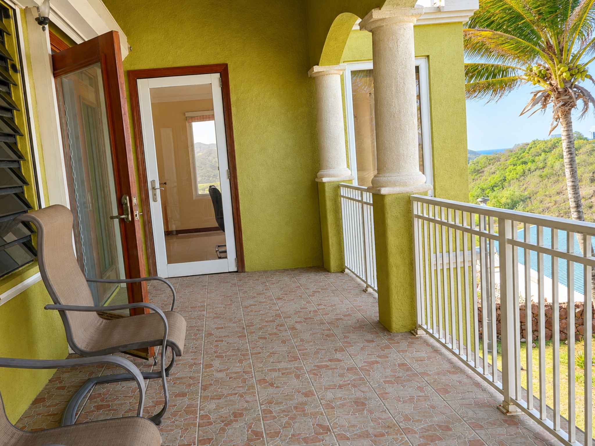 Tiled balcony with white railings and columns, overlooking palms and green hillside