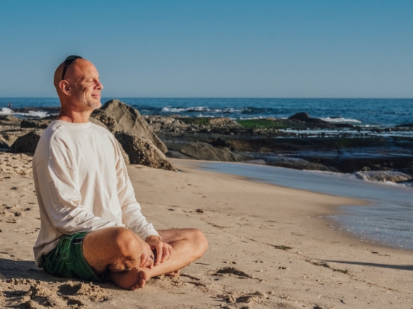 bald man smiling while sitting in lotus pose on the beach
